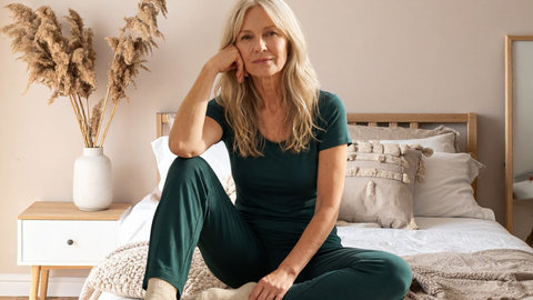 A woman with long silver-blonde hair sits on the edge of her made bed in a deep green A DOMANI cooling sleep set, rested and dry after a full night.